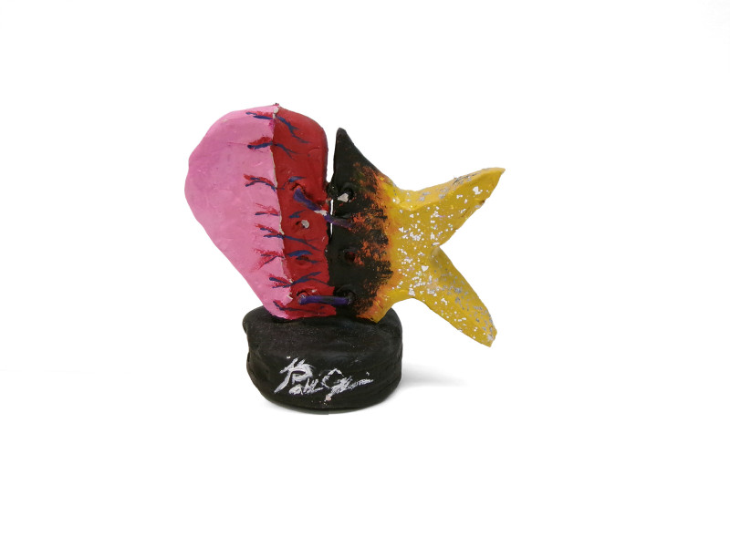 A tabletop sculpture that looks like a fish on a black pedestal. The front half of the fish looks like a heart, with some veins painted in. The back half is more classic, with a body in black and tail fins in yellow and orange. The pedestal is signed in silver.