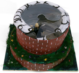 A sculpture of a physical enclosure and a small person. The enclosure is made of two circular tiers. The walls of the tiers are reddish brown with white details. The ground, and the horizontal area above the first tier, is covered in grass and what look like flowers. At the top, there is a piece of clear, cracked plastic. The edges of the plastic are painted white, with black numerals representing the face of a clock. A bald man with a beard is within the structure. He has one arm lifted and he looks toward the viewer.