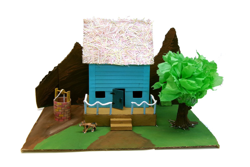A detailed sculpture of a house and its yard made mostly from paper. The house is blue, with defined siding and a confetti-like roof. There is a porch, its railing is made from string. There are mountain shapes behind the house, a well to its left, and a tree to its right. There is a path and two steps leading to the porch, and a dog is in the yard.