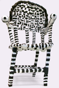 A fanciful chair sculpture painted with black and white stripes. The pommels of the arm rests look like eyeballs. The seat back is woven. The entire thing is a little off-kilter.