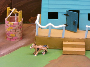 A detail photograph of the previous sculpture. This view shows the dog, well, porch, railing, and the front door of the house from close up.